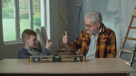 Photo for Grandpa and grandson are sitting at the table near the water level and showing a thumbs up gesture. An elderly man and a teenager pose against the background of a room with a window and a ladder. The - Royalty Free Image