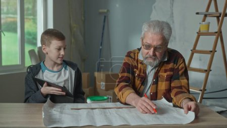 Photo for Grandpa is planning repairs and making notes on project with pencil and ruler. Grandson helps granddad solve complex calculation using digital tablet. An elderly man and a young guy are sitting at a - Royalty Free Image