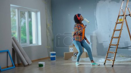Téléchargez les photos : Young african american woman in big wireless red headphones is enjoying music. Black female in checkered shirt sings into paint roller as if into microphone and dances. Ladder, cardboard boxes - en image libre de droit