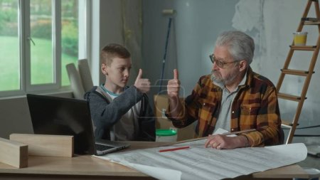 Foto de Grandpa plans renovations using a paper apartment project. Grandson helps granddad solve complex calculation using a laptop. Elderly man and young guy are sitting against the window and show thumbs up - Imagen libre de derechos