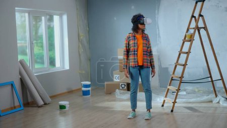 Téléchargez les photos : Young african american woman in virtual reality headset visualizing interior of room. Female in checkered shirt makes choice using virtual technology. Concept of modern apartment renovation, finishing - en image libre de droit