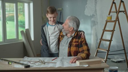 Foto de Grandson friendly is hugging his grandfather. Elderly man and young guy are smiling together against the background of the window, ladders and cardboard boxes. The concept of family, joint renovation - Imagen libre de derechos