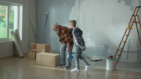 Photo for The elderly man cries out in pain in his back and young guy frightenedly rushes to his aid. Grandpa and grandson pose in the apartment against the backdrop of a ladder, cardboard boxes, paint cans and - Royalty Free Image