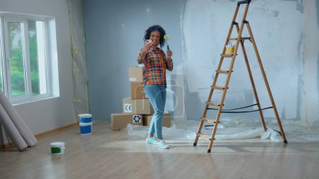 Photo for Young African American woman is making video call using her mobile phone, smiling and showing a paint roller. Black female in checkered shirt communicates remotely through camera. Concept of repair - Royalty Free Image