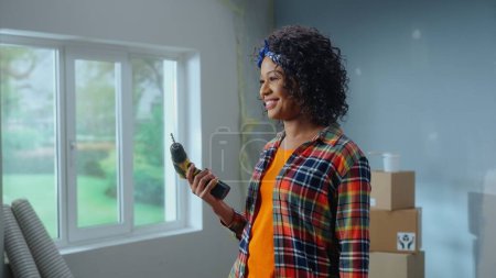 Téléchargez les photos : Young African American woman is turning on an electric screwdriver and smiling. Black female standing against the background of cardboard boxes and window. Concept of apartment renovation - en image libre de droit