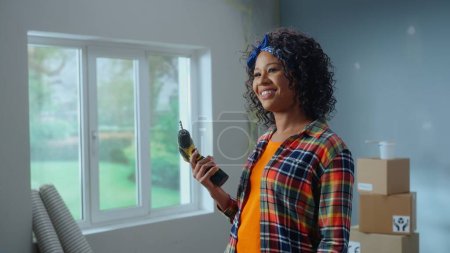Photo for Young African American woman is turning on an electric screwdriver and smiling. Black female standing against the background of cardboard boxes and window. Concept of apartment renovation - Royalty Free Image