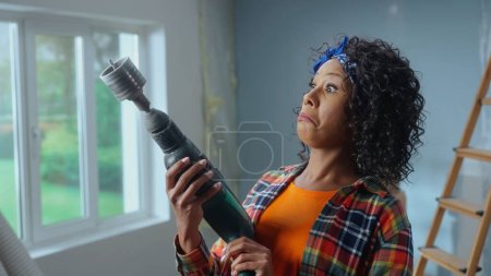 Photo for Young African American woman turns on an electric perforator with nozzle for drilling sockets. Black female look at drill with surprise against backdrop of an apartment in the process of being - Royalty Free Image