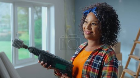 Foto de A young African American woman holds an electric hammer drill with a nozzle for drilling holes in her hands. A black woman poses in front of a window and smiles. The concept of repair in the apartment - Imagen libre de derechos
