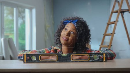 Téléchargez les photos : Une jeune Afro-Américaine pose à côté d'un niveau de bulle sur une table. Une femme noire pense à quelque chose dans le contexte d'un appartement en cours de rénovation. Le concept de réparation - en image libre de droit