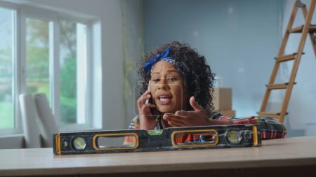 Photo for A young African American woman poses next to a building bubble level on a table. A black woman calls the master on the phone and asks for help in repairing the apartment - Royalty Free Image