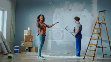 Photo for African American woman is giving directions to male house painter painting wall with white paint using long paint roller. Repairman in blue construction overalls is doing repairs in an apartment - Royalty Free Image