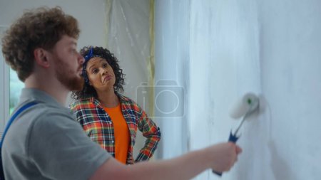 Photo for African American woman is watching a male painter work who paints a wall with white paint using a paint roller. Repairman in blue construction suit is doing repairs in an apartment. Concept of repair - Royalty Free Image