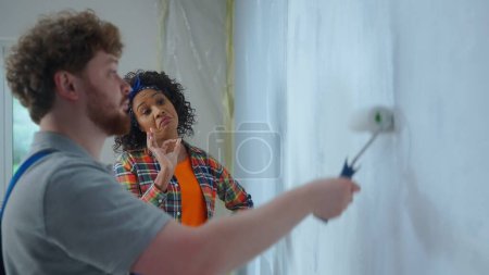 Photo for African American woman is showing ok gesture to a male house painter painting a wall with white paint using a paint roller. Repairman in blue construction suit is doing repairs in an apartment - Royalty Free Image