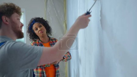 Photo for African American woman is watching a male painter work who paints a wall with white paint using a paint roller. Repairman in blue construction suit is doing repairs in an apartment. Concept of repair - Royalty Free Image