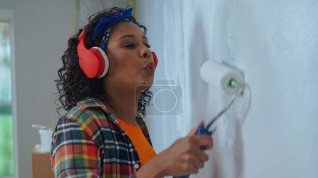 Téléchargez les photos : African American woman in red headphones paints wall with white paint using paint roller and enjoy the music. Black female making repairs in an apartment. Concept of repair, finishing works, interior - en image libre de droit