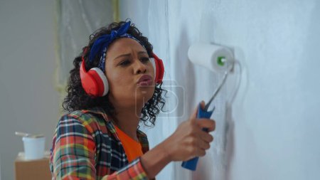 Photo for African American woman in red headphones paints wall with white paint using paint roller and enjoy the music. Black female making repairs in an apartment. Concept of repair, finishing works, interior - Royalty Free Image