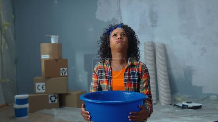 Téléchargez les photos : Femme afro-américaine bouleversée tenant bol bleu dans lequel gouttes d'eau coulent de fuite de plafond. Une femme noire pose devant des boîtes en carton, des rouleaux de papier peint et un mur peint. Appartement - en image libre de droit