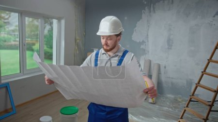 Photo for Male foreman is looking through large sheet with an apartment plan and thinking over repair project in flat. Redhead man with beard in blue construction overalls and white helmet is planning finishing - Royalty Free Image