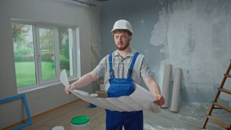 Photo for Male foreman is looking through large sheet with an apartment plan and thinking over repair project. Redhead man with beard in blue construction overalls and white helmet looks in disbelief - Royalty Free Image