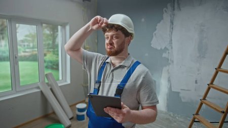 Photo for Male foreman in blue construction overalls and white helmet is planning renovation project in an apartment using tablet. Redhead master man with beard thinking, viewing and visualizing ideas against - Royalty Free Image