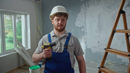 Photo for Male foreman in blue overalls and white helmet is planning renovation project and turning on an electric screwdriver. Redhead master man thinking something against background of an apartment in - Royalty Free Image