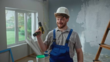 Foto de Male foreman in blue overalls and white helmet is planning renovation project and turning on an electric screwdriver. Redhead master man thinking something against background of an apartment in - Imagen libre de derechos