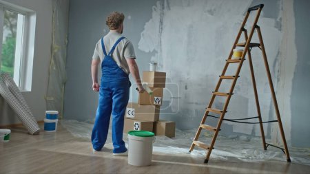 Photo for Rear view of a foreman or house painter in blue construction overalls. A red haired man with a paint roller in his hands plans to paint the walls in the apartment. The concept of repair, finishing - Royalty Free Image