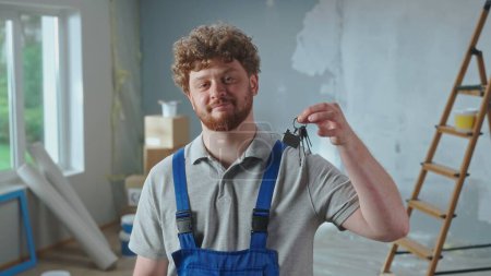 Photo for Repairman worker in blue construction overalls is showing bunch of keys. Portrait of redhead man is posing against backdrop of apartment in process of renovation, ladder, cardboard boxes, window - Royalty Free Image