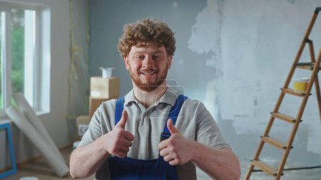 Photo for Repairman worker in blue construction overalls is smiling and showing thumbs up. Portrait of redhead man is showing gesture of recommendation and posing against backdrop of apartment in process of - Royalty Free Image