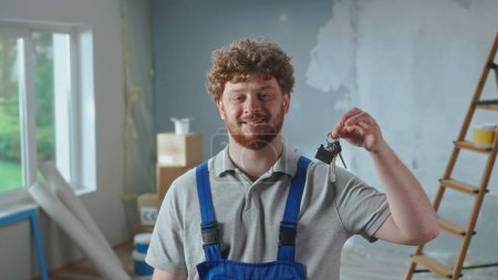 Photo for Repairman worker in blue construction overalls showing bunch of keys and smiling. Portrait of redhead man posing against backdrop of apartment in process of renovation, ladder, cardboard boxes, window - Royalty Free Image