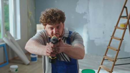 Téléchargez les photos : A repairman in blue construction overalls holds an electric drill in his hands and points it straight at the camera. Portrait of a red haired man posing against the backdrop of an apartment with a - en image libre de droit