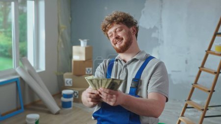 Téléchargez les photos : Travailleur réparateur en salopette bleue compte l'argent et sourit. Portrait d'un homme roux pose sur fond d'appartement, échelle, boîtes en carton, fenêtre. Concept de réparation, travaux de finition - en image libre de droit