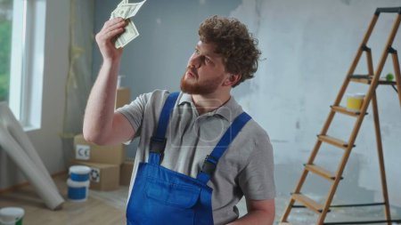 Photo for A repairman in blue overalls examines paper banknotes. Portrait of a red haired man posing against the backdrop of an apartment, ladder, cardboard boxes, windows. The concept of repair, finishing work - Royalty Free Image