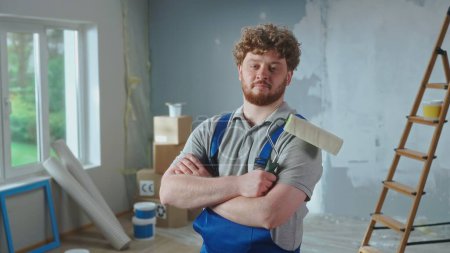 Photo for Repairman in blue overalls is looking confidently at camera and crossing arms with paint roller. Portrait of redhead man is posing against backdrop of renovation apartment, ladder, cardboard boxes - Royalty Free Image