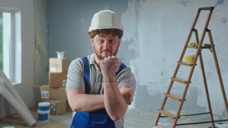 Photo for Repairman worker in blue overalls looking at camera and showing obscene gesture, fuck you. Portrait of redhead man posing against backdrop of renovation apartment, ladder, cardboard boxes, window - Royalty Free Image
