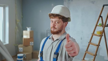Téléchargez les photos : Repairman worker in blue overalls and white helmet is smiling and showing thumbs up. Portrait of redhead man is showing gesture of like and posing against backdrop of apartment in process of - en image libre de droit