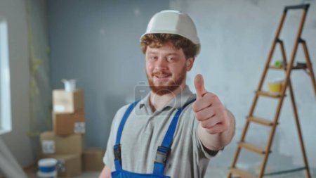 Photo for Repairman worker in blue overalls and white helmet is smiling and showing thumbs up. Portrait of redhead man is showing gesture of like and posing against backdrop of apartment in process of - Royalty Free Image