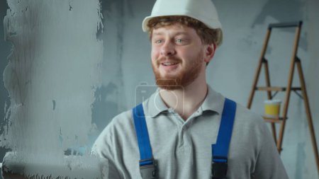 Photo for Repairman worker in blue overalls and white helmet is painting glass with white paint using roller. Redhead man is filling the screen frame with white paint against backdrop of renovation apartment - Royalty Free Image