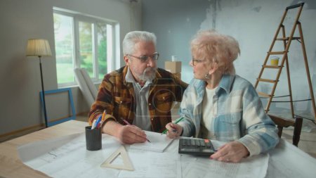 Téléchargez les photos : Elderly man and woman calculating expenses of repairing an apartment on calculator. Aged couple sitting at a table and planning the improvement of their home. Concept of repair, decoration, interior - en image libre de droit