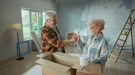 Téléchargez les photos : Homme et femme âgés sortent le pinceau d'une boîte en carton et sourient. Un couple âgé planifie des réparations dans un appartement et discute de la peinture des murs. Concept de réparation, décoration - en image libre de droit