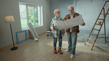 Téléchargez les photos : Elderly man and woman are looking through sheet with plan of an apartment and discussing renovation project. Aged couple is planning improvement of their home. Ladder, cardboard boxes and window in - en image libre de droit