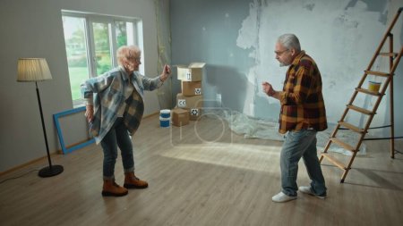 Téléchargez les photos : Homme et femme âgés dansent gaiement et sourient. Deux retraités s'amusent et célèbrent la crémaillère. Échelle, boîtes en carton et fenêtre encadrée. Le concept de réparation, décoration, intérieur - en image libre de droit