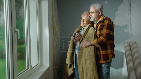 Foto de Un anciano abraza a una anciana envuelta en una tela a cuadros y con una taza en las manos. Un par de pensionistas están soñando con algo mirando por la ventana y sonriendo felizmente. Ambiente acogedor en el hogar - Imagen libre de derechos