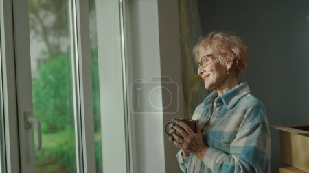 Photo for An elderly woman with a cup in her hands looks out the window, thinks about something pleasant and smiles. An aged woman poses at the window against the backdrop of repairs, cardboard boxes and - Royalty Free Image