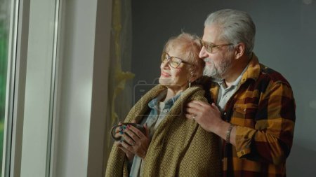 Photo for An elderly man hugs an aged woman wrapped in a plaid and with a cup in her hands. A couple of pensioners are dreaming about something looking out the window and smiling happily. Cozy home environment - Royalty Free Image