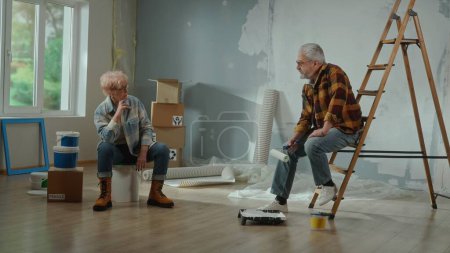 Foto de An upset elderly man with a roller in his hands sits on the stairs. An elderly woman sitting on a large can of paint and looks tiredly at a man. A tired couple of pensioners are resting during the - Imagen libre de derechos