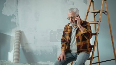 Téléchargez les photos : Tired elderly man is sitting on a ladder and calling a master to repair an apartment using a mobile phone. Aged pensioner solved problem with help of the master. Concept of repair, decoration - en image libre de droit