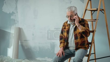 Foto de El anciano cansado está sentado en una escalera y llamando a un maestro para reparar un apartamento usando un teléfono móvil. Pensionista de edad resuelto el problema con la ayuda del maestro. Concepto de reparación, decoración - Imagen libre de derechos