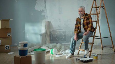 Téléchargez les photos : A tired elderly man sits on a ladder and thinks about something. An elderly pensioner poses in front of an apartment with cardboard boxes, wallpaper and building materials - en image libre de droit