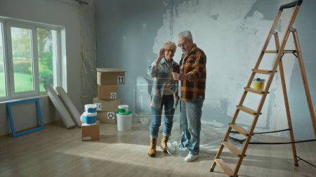 Téléchargez les photos : Elderly man and woman are talking on a video call using their mobile phone. Aged couple communicates with someone and showing repairs in the apartment, ladder, cardboard boxes, a window and wallpaper - en image libre de droit